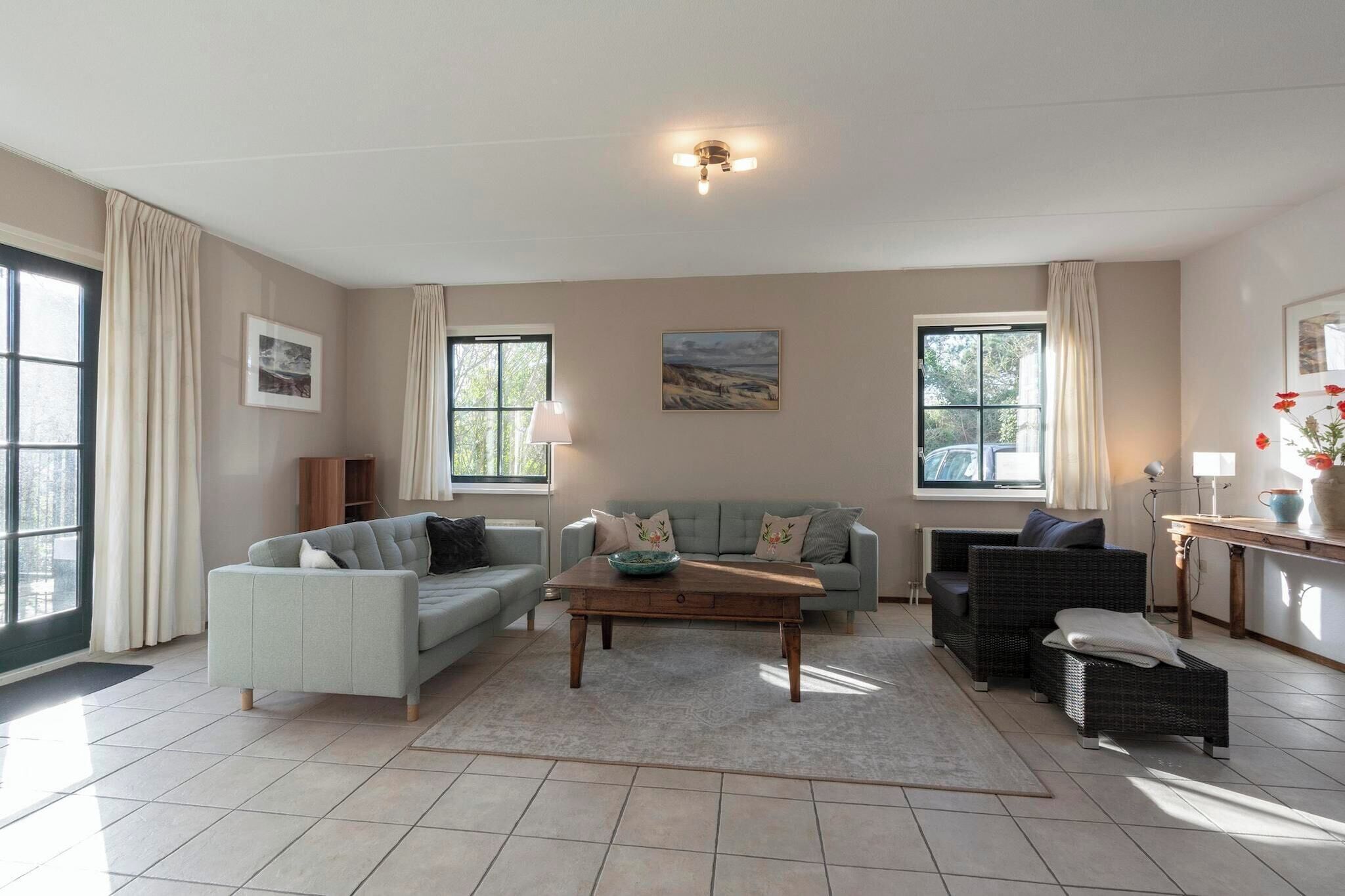 A spacious living room with a light grey sofa, a wooden coffee table, and a wicker sofa. There are two windows with curtains, a painting on the wall, and a lamp on a side table.