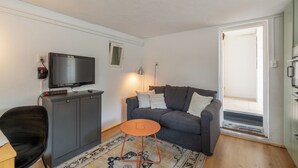 A living room with a grey sofa, a small orange table, a black chair, a TV on a grey cabinet, and a patterned rug. - Holiday Home in Callantsoog near Beach (Callantsoog)