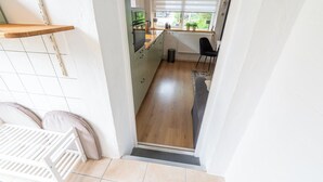 A narrow hallway leading to a kitchen with a radiator, a window with blinds, and a black chair. - Holiday Home in Callantsoog near Beach (Callantsoog)