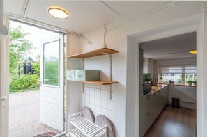 A kitchen with white cabinets, a wooden shelf, and a view of the garden. - Holiday Home in Callantsoog near Beach (Callantsoog)