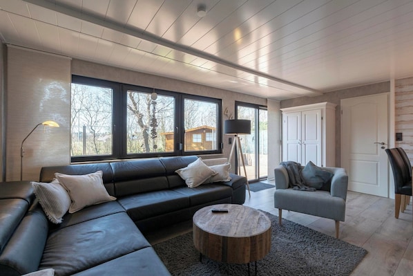 Una sala de estar moderna con un sofá modular, una mesa redonda de centro y un sillón gris. Amplias ventanas ofrecen luz natural y vistas al exterior.