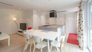 A modern kitchen with white cabinets, a central island, and a built-in stove. There is a dining area with a white table and chairs. - Duinland 146 for 4 guests with garden (Sint Maartenszee)