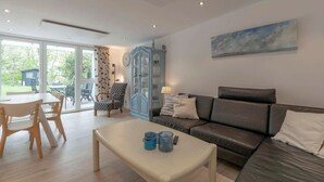 A modern living room with a leather sofa, a coffee table with candles, a dining area with a table and chairs, and a large window with a view of the outdoors. - Duinland 146 for 4 guests with garden (Sint Maartenszee)