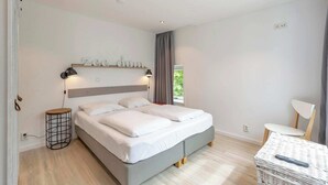 A modern bedroom with a bed, bedside lamps, a small round table, a chair, and a window with curtains. - Duinland 146 for 4 guests with garden (Sint Maartenszee)