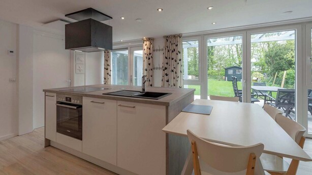 A modern kitchen with a built-in oven, a large island with a sink, and a dining area with a table and chairs. - Duinland 146 for 4 guests with garden (Sint Maartenszee)