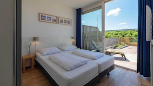 Ein Schlafzimmer mit zwei Betten, einem kleinen Nachttisch, einer Lampe und einem Fenster mit Blick auf einen Balkon und Grünflächen.