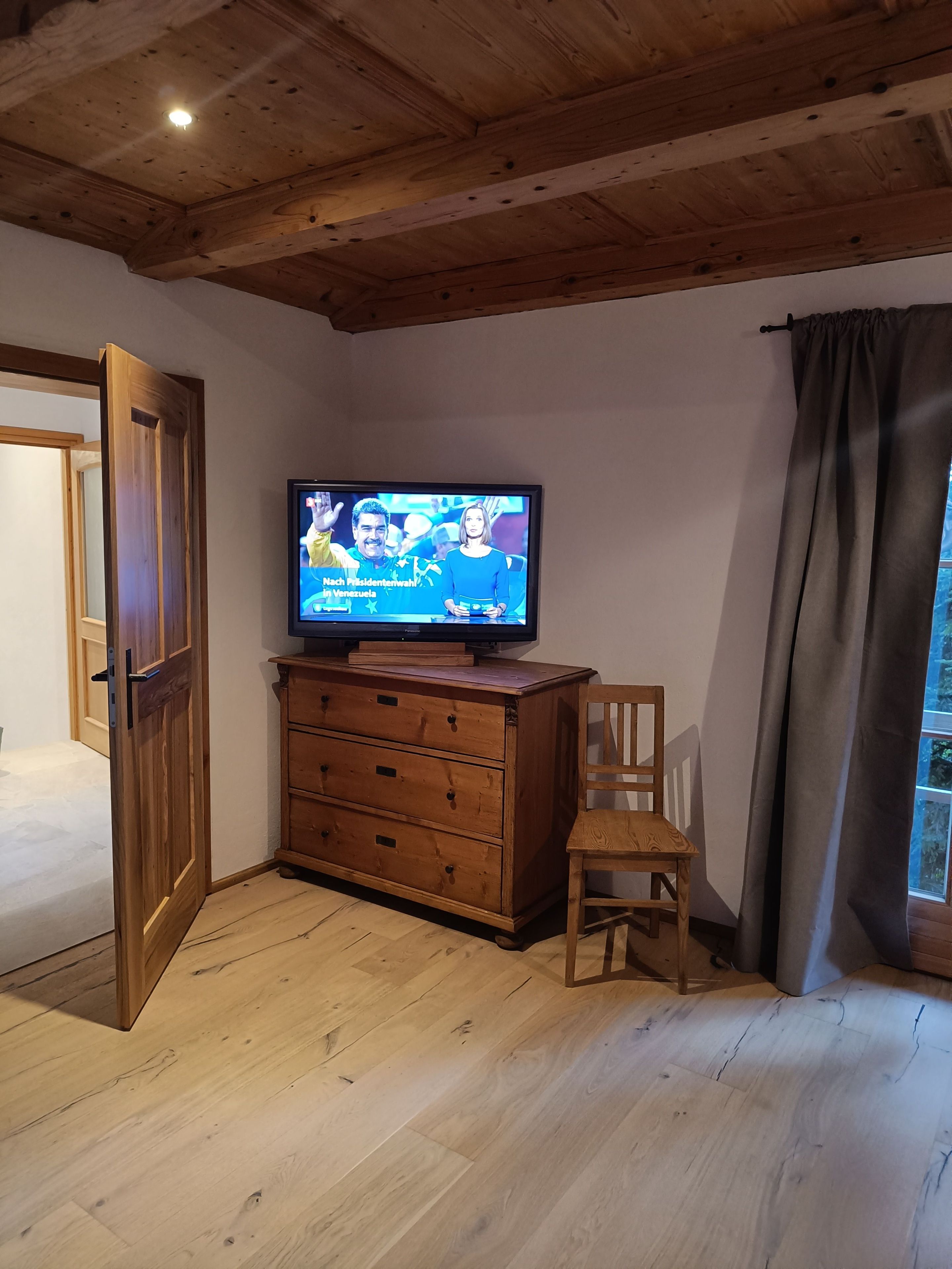 Ein Zimmer mit Holzboden, einem Fernseher auf einer Holzkommode, einem Holzstuhl und einem Fenster mit Vorhängen.