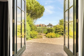 Una puerta abierta que da a un jardín con un sendero de grava, rodeado de follaje y una hilera de casas con tejados de paja.