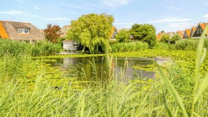 A tranquil riverside scene with a small building, lush greenery, and traditional houses in the background. - Villa with view over the park De Buitenplaats (Callantsoog)