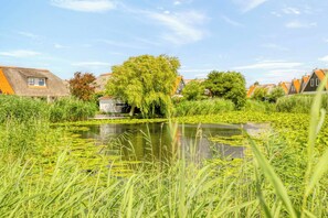 A tranquil riverside scene with a small building, lush greenery, and traditional houses in the background. - Villa with view over the park De Buitenplaats (Callantsoog)