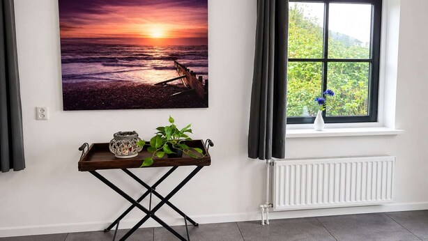A room with a large sunset painting, a window with curtains, a radiator, and a small table with a plant and a candle. - Villa with view over the park De Buitenplaats (Callantsoog)