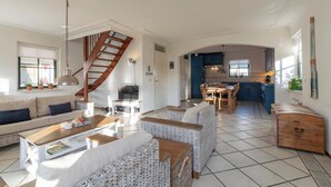 A spacious living area with a sofa, wicker furniture, a staircase, and a kitchen in the background. - De Buitenplaats 7 villa with dune views (Callantsoog)