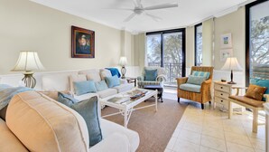 A spacious living room with a white ceiling fan, a large window offering a view of trees, a beige sofa with blue cushions, a wicker armchair, and a white coffee table. - 123D Shipwatch by AvantStay | Beach Access + Pool (Isle Of Palms)