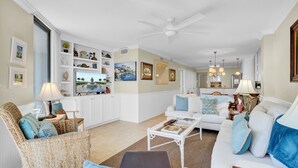 A spacious living room with a white sofa set, a wicker chair, a coffee table, and a television. - 123D Shipwatch by AvantStay | Beach Access + Pool (Isle Of Palms)