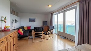A modern living room with a sofa, a wooden cabinet, a TV, and a balcony with a view. - Cozy Stay Near Dunes (Julianadorp aan Zee)