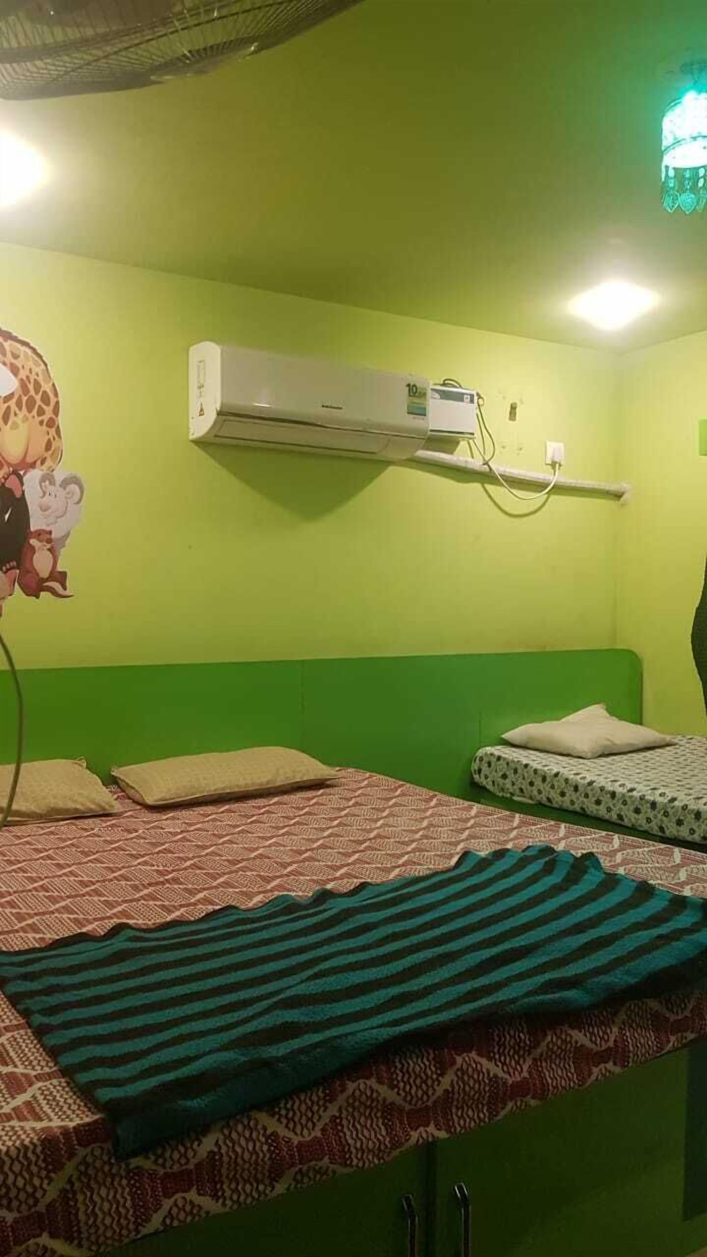 Une pièce avec un mur vert, un lit avec une literie à motifs, un climatiseur fixé au mur et un luminaire au plafond.