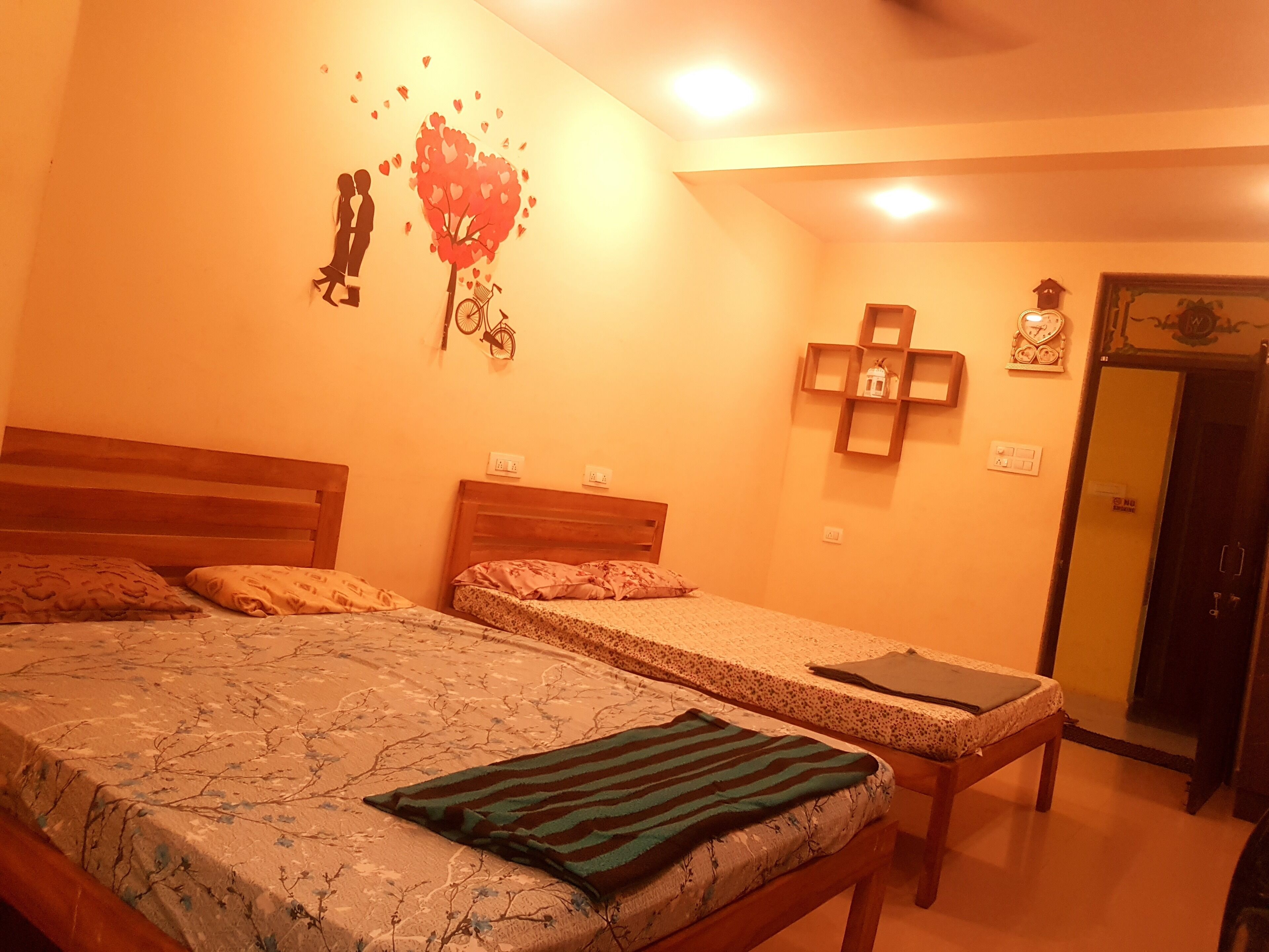 A room with two single beds, a wall clock, a small shelf with decorative items, and a door with a handle.