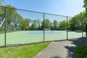 Una cancha de tenis rodeada por una cerca de malla metálica, con un camino pavimentado que la conduce.