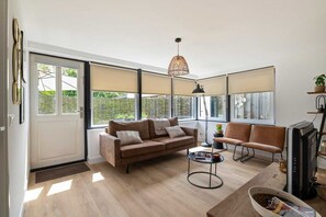 A modern living room with a sofa, armchair, and coffee table, large windows with blinds, and a hanging light fixture.