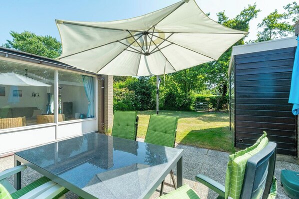 Una mesa con cubierta de vidrio, una sombrilla grande, sillas con cojines verdes y una vista a un jardín y una casa al fondo.