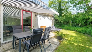 A patio area with a table and chairs, a covered seating area, and a view of a lush garden. - Holiday Home near Dutch Beach with Pool (Sint Maartenszee)