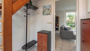A staircase with a wooden handrail, a black coat rack, a small wooden cabinet, and a grey sofa in a living area with a window and a painting on the wall. - Holiday Home near Dutch Beach with Pool (Sint Maartenszee)