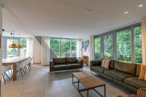 A spacious living area with a sofa, a coffee table, and a dining table with chairs. Large windows provide a view of greenery outside.