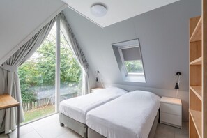A bedroom with two beds, a desk, a nightstand, a mirror, and a window with a view of greenery.