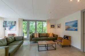 Una sala de estar con un sofá, una mesa de centro y una silla de mimbre. Hay ventanas grandes con cortinas, un cuadro en la pared y una puerta.