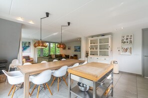 Ein moderner Essbereich mit Holztisch und weißen Stühlen, eine Küche mit weißen Schränken und Blick auf Grünflächen.