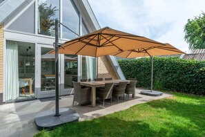 Ein moderner Essbereich im Freien mit Tisch und Stühlen, einem großen Sonnenschirm und einer Glastür zu einem Innenraum.
