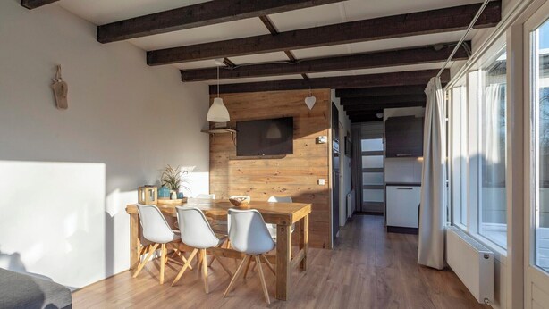 A modern kitchen with wooden beams, a dining area with a wooden table and chairs, and a view of the outside through large windows. - Duinland 245 bungalow for 6 guests near sea (Sint Maartenszee)