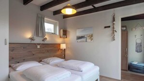 A bedroom with a wooden headboard, two beds with white linens, a window with a curtain, a wall-mounted lamp, a beach-themed picture, and a door leading to another room. - Duinland 245 bungalow for 6 guests near sea (Sint Maartenszee)