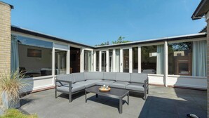 A modern outdoor patio with a grey sectional sofa, a black coffee table, and a glass door leading to a covered area. - Duinland 245 bungalow for 6 guests near sea (Sint Maartenszee)