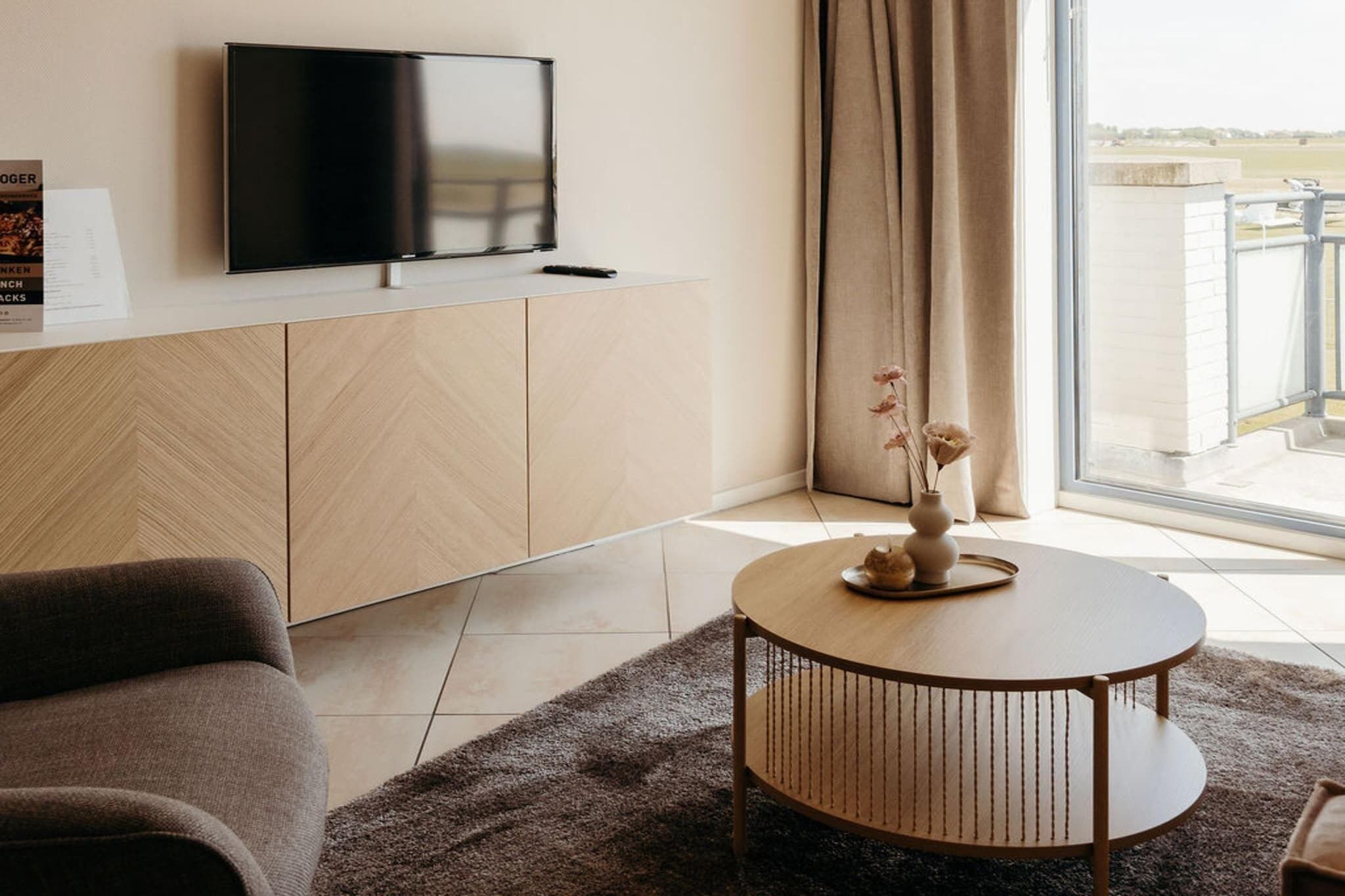 Ein modernes Wohnzimmer mit einem an einer Holzeinrichtung befestigten Flachbildfernseher, einem runden Couchtisch mit einer Vase und Blick auf einen Balkon durch große Fenster.