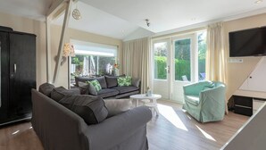 En moderne stue med sofa, lænestol og et lille bord. Der er et TV ophængt på væggen og et vindue med gardiner.