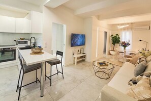 Una cocina moderna con armarios blancos, un comedor con mesa y sillas blancas, una sala de estar con sofá y mesa baja, y un televisor montado en la pared.