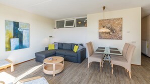A modern living room with a grey sectional sofa, a round coffee table, and a dining area with a glass table and chairs. - Apartment 189 in Julianadorp aan Zee (Julianadorp aan Zee)