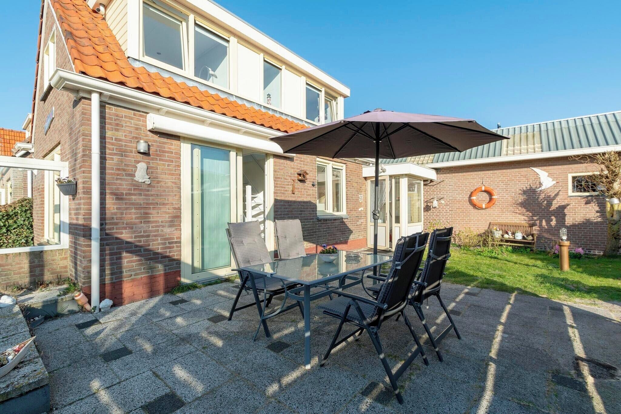 Eine Terrasse mit Glastisch und Stühlen, einem großen Sonnenschirm und einem Ziegelhaus mit Ziegeldach.