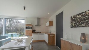 A modern kitchen with white cabinets, a wooden countertop, and a built-in oven. There is a dining area with a white table and chairs, and a large window offering a view of the outdoors. - Bungalow near Dutch Beach and Bulb Fields (Sint Maartensvlotbrug)