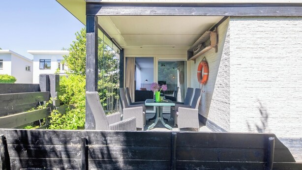 Un espacio moderno al aire libre con mobiliario de mimbre y una mesa, con vistas a un jardín y un edificio de fachada blanca.