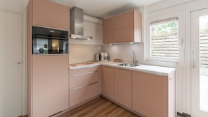 A modern kitchen with pink cabinets, a stainless steel range hood, a built-in oven, a coffee maker, and a window with blinds. - Holiday home Effe Sprage in Callanstoog Centre (Callantsoog)