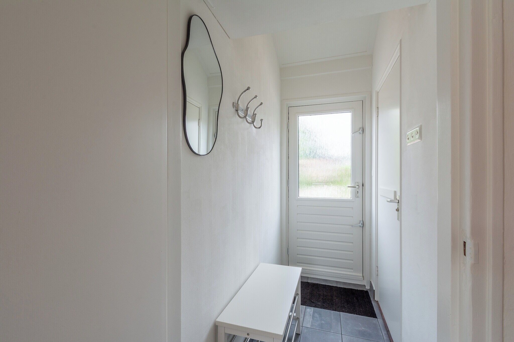 Un pasillo estrecho con una puerta blanca, un banco blanco y un espejo fijado a la pared.