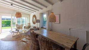 A spacious living area with a wooden dining table, wicker chairs, and a large window offering a view of greenery. - Bungalow in Callantsoog near the Beach (Callantsoog)