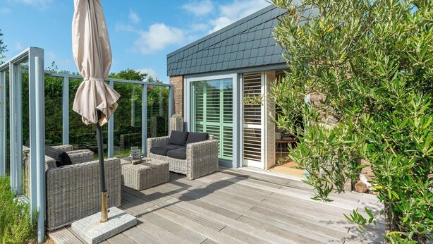 A patio with wicker furniture and a covered umbrella, a glass door, and a view of olive trees. - Bungalow in Callantsoog near the Beach (Callantsoog)
