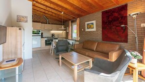 A cozy living room with a wooden ceiling, a sofa, a coffee table, and a kitchen in the background. - Bungalow near Beach and Forest in De Blenck (Callantsoog)