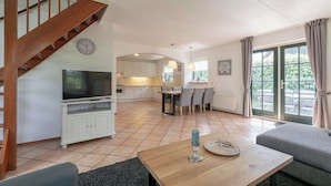 A spacious living area with a wooden staircase, a dining table with chairs, a television, and a kitchen in the background.