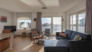 A modern living room with a sofa, a dining table, and a TV. There is a large window with a view of buildings and a sailboat model.