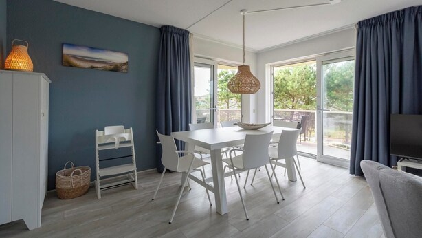 A modern kitchen with white cabinetry, a dining table, and chairs. There is a large window with curtains, a hanging light fixture, and a television on a stand.