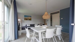 A modern dining area with a white table and chairs, a hanging wicker light fixture, and a view of the outdoors through large windows.